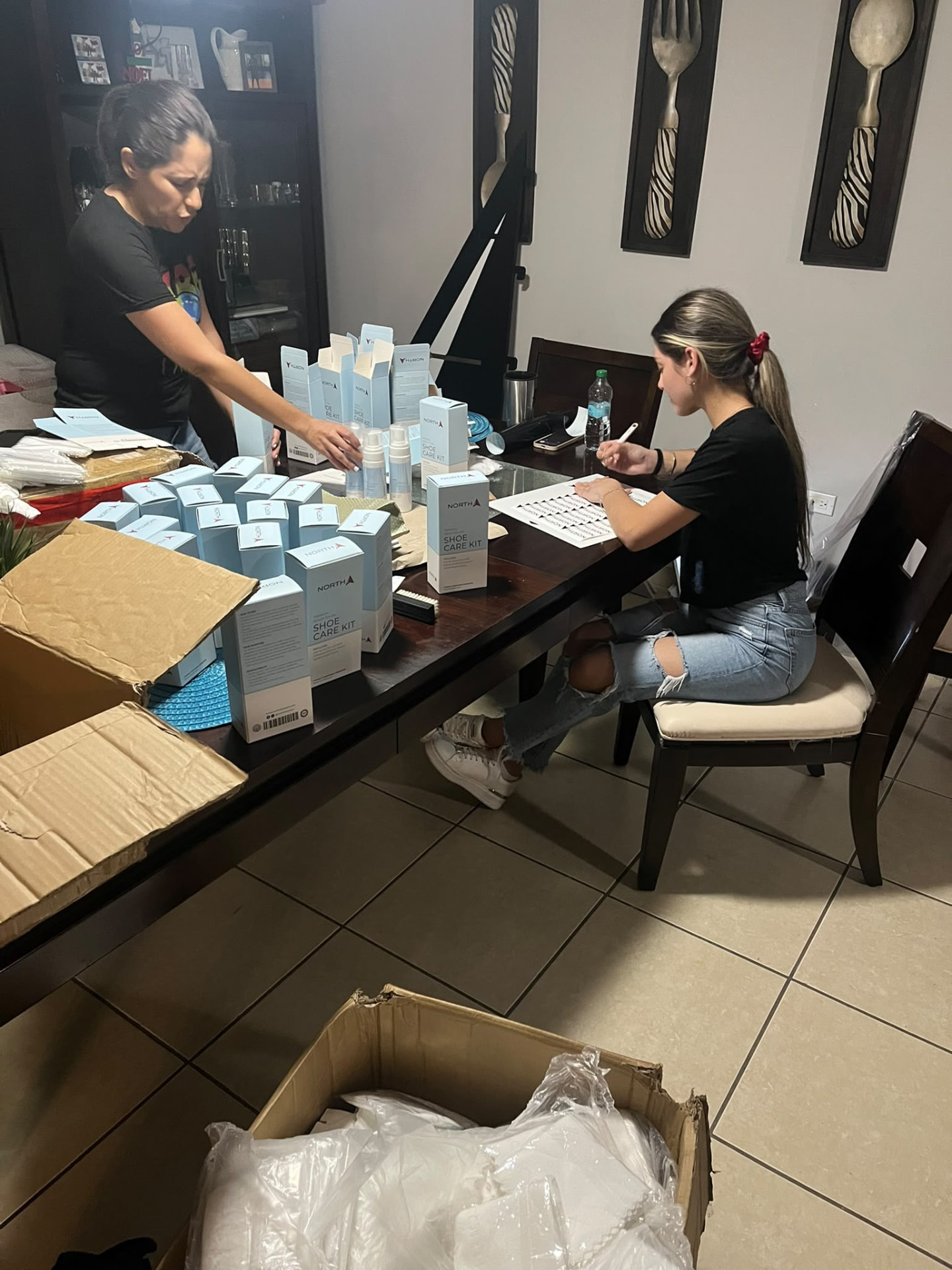 Two people packing NORTH Shoe Care Kit boxes at a home dining table